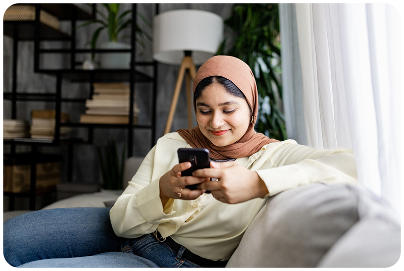 A woman in a hijab on a sofa, looking relieved at her phone