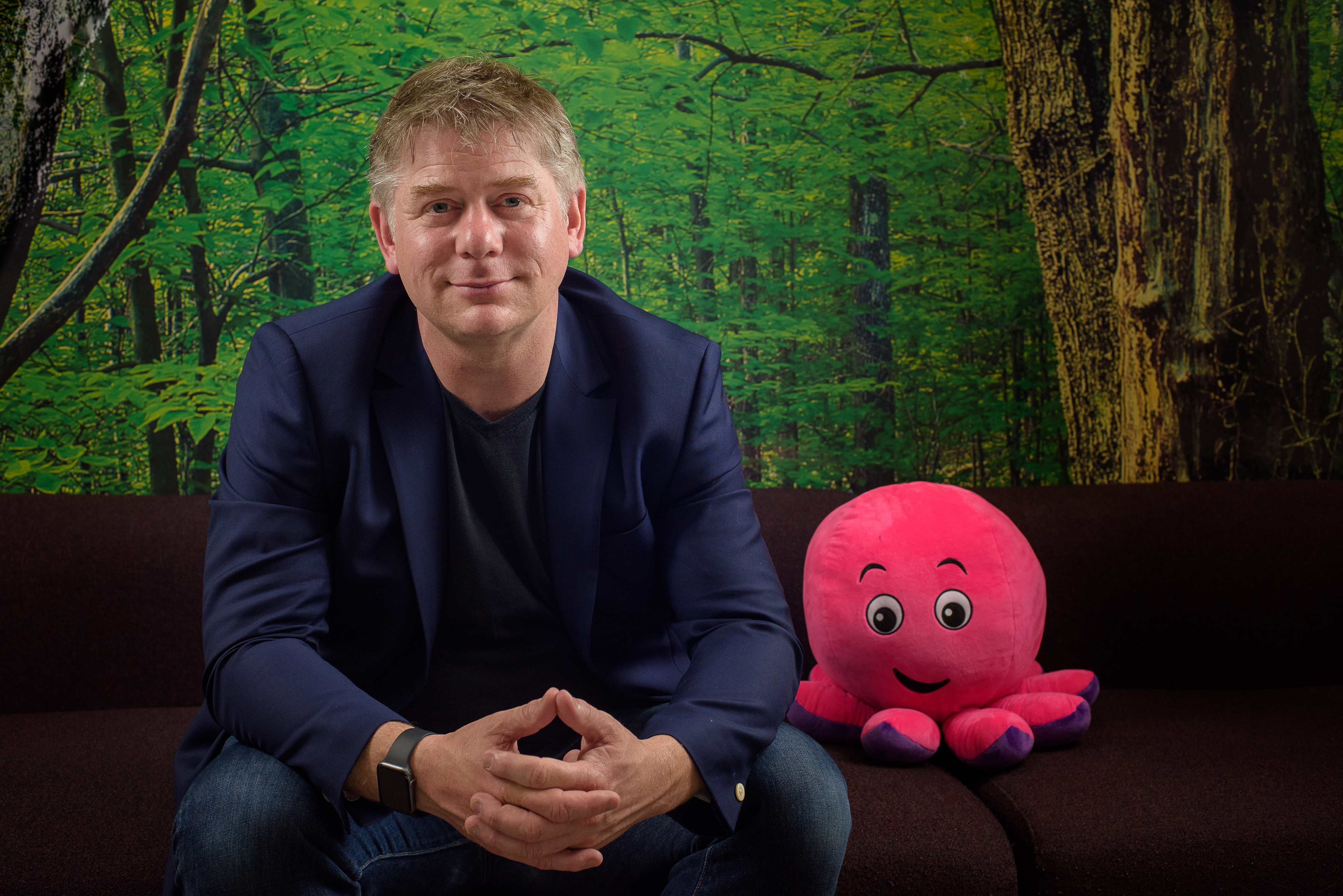 An image of Greg sitting on a sofa next to a pink cuddly octopus toy